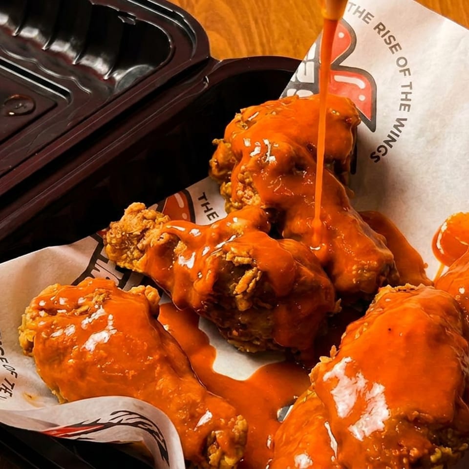 Orange sauce pouring over fried chicken wings in a branded takeout container.