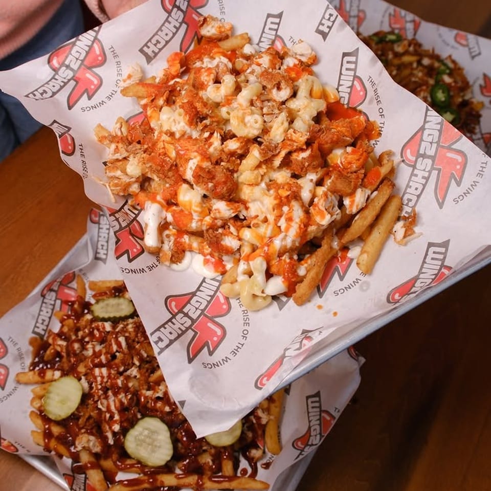 Loaded fries topped with crispy chicken, macaroni, cheese, and orange and white sauces, with another pickle-topped serving below.