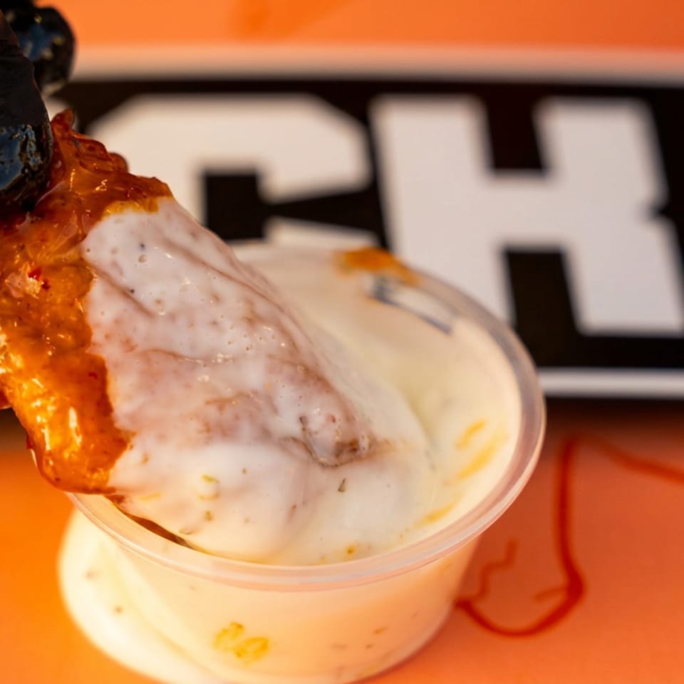 Hand in black glove dipping a saucy chicken wing into a white dipping sauce cup.